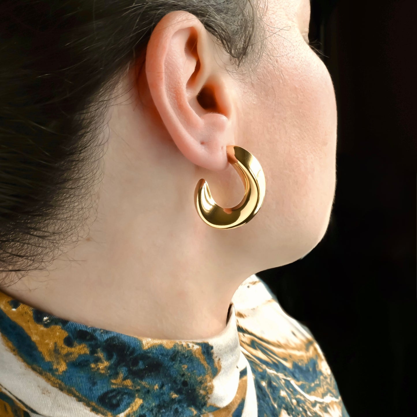 Side view of chunky gold hoop earrings worn by a person with a dark background