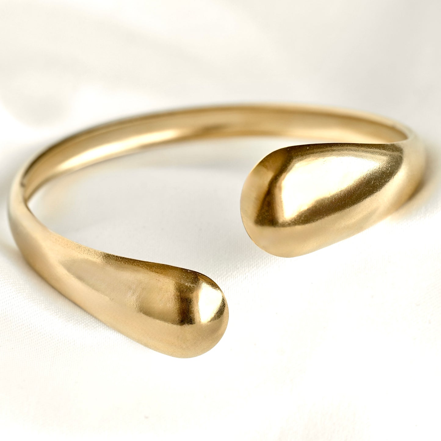 A polished gold-tone open cuff bracelet with a modern, organic teardrop design, photographed on a clean white fabric background.
