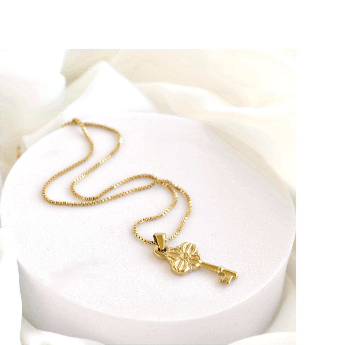A gold key pendant necklace on a delicate gold chain, displayed on a white cylindrical pedestal with soft white fabric in the background.