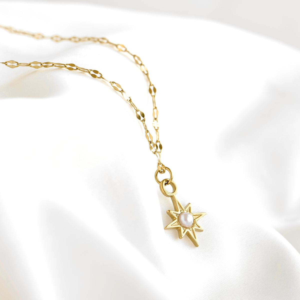A gold chain necklace with a starburst pendant featuring a central white pearl, displayed on a soft white satin fabric background.