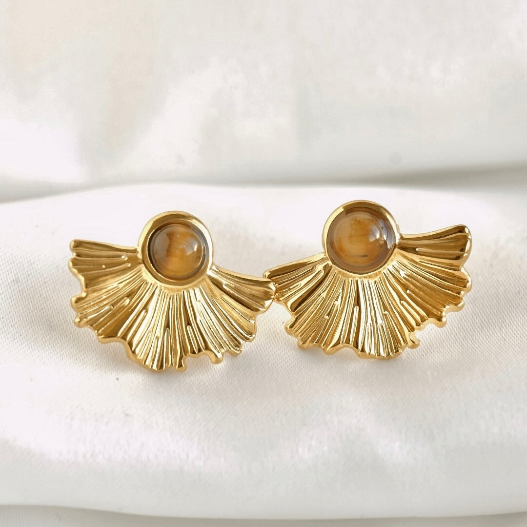 A pair of golden fan-shaped drop earrings with oval brown tiger's eye stones, displayed on a soft white fabric background.