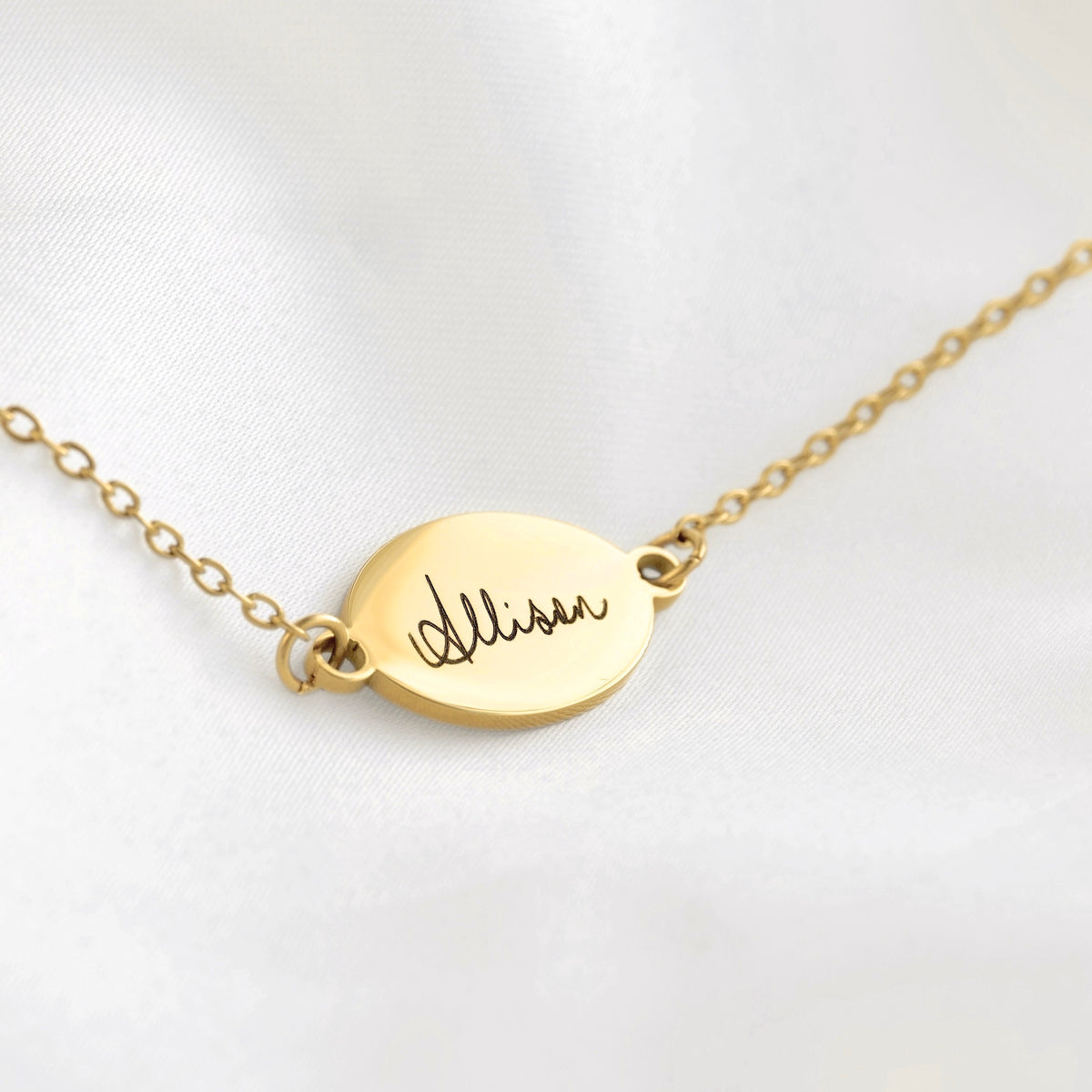 A gold oval pendant necklace engraved with the name 'Allison' in cursive, resting on a white satin fabric background.