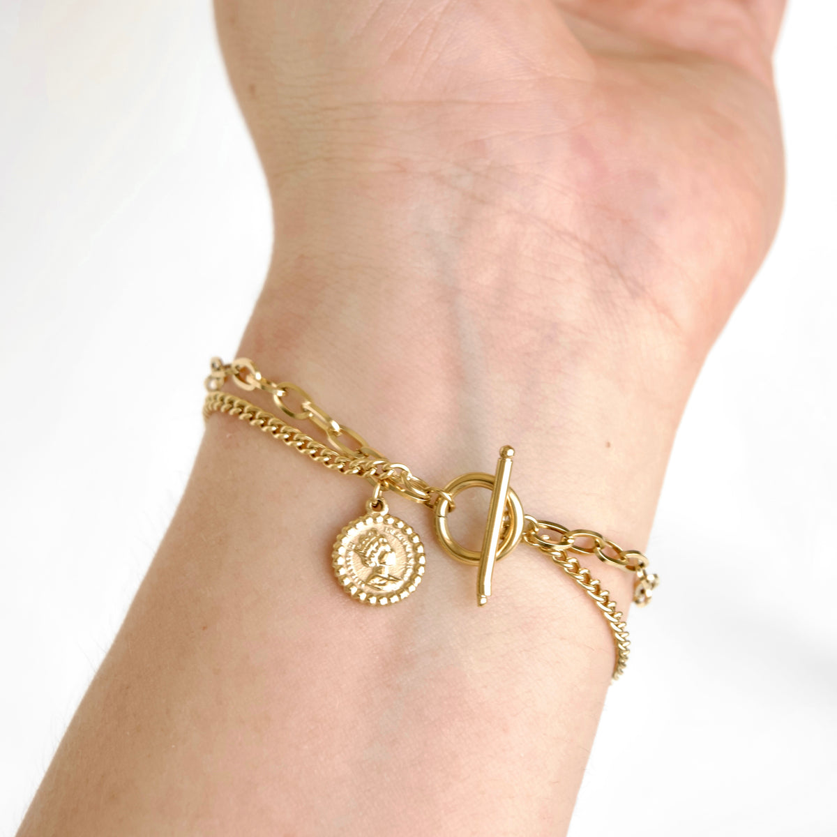 A delicate gold chain bracelet with a toggle clasp and a small circular coin charm, shown worn on a person's wrist against a clean white background.