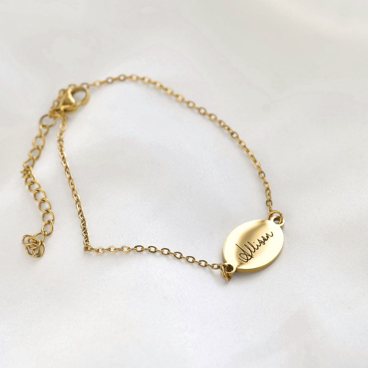 A delicate gold chain bracelet with an oval engraved nameplate and adjustable lobster clasp, displayed on a soft white fabric background.