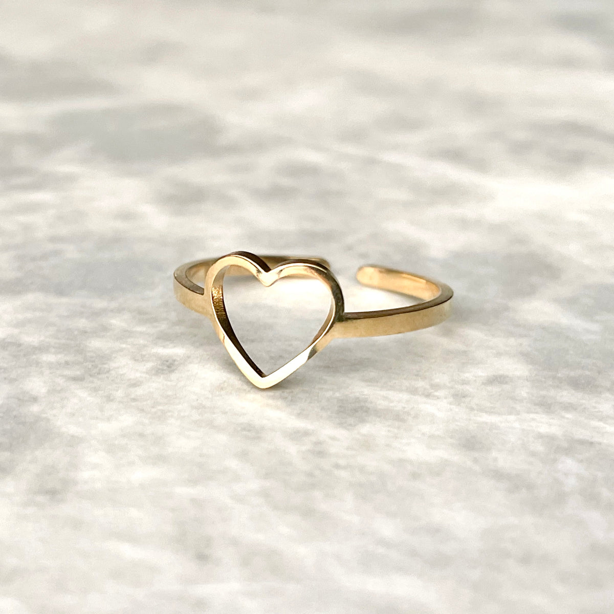 Angled view of adjustable gold ring with an open heart charm in front on a textured gray background