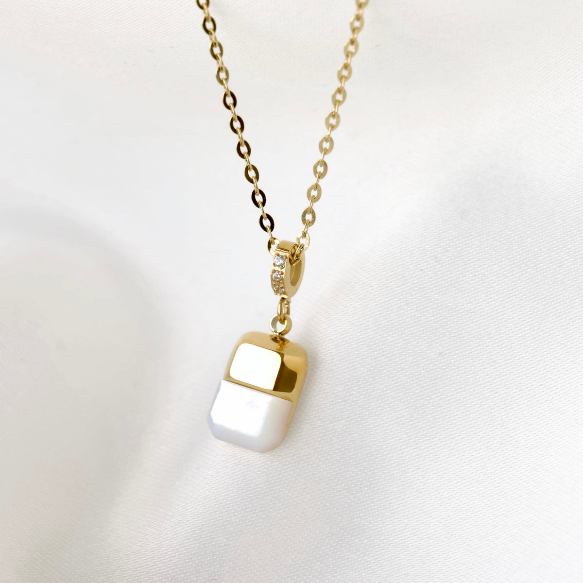 A gold chain necklace with a small rectangular pendant featuring a glossy white pearl top bottom and a polished gold top half and a pave CZ pendant bail, displayed on a soft white fabric background.