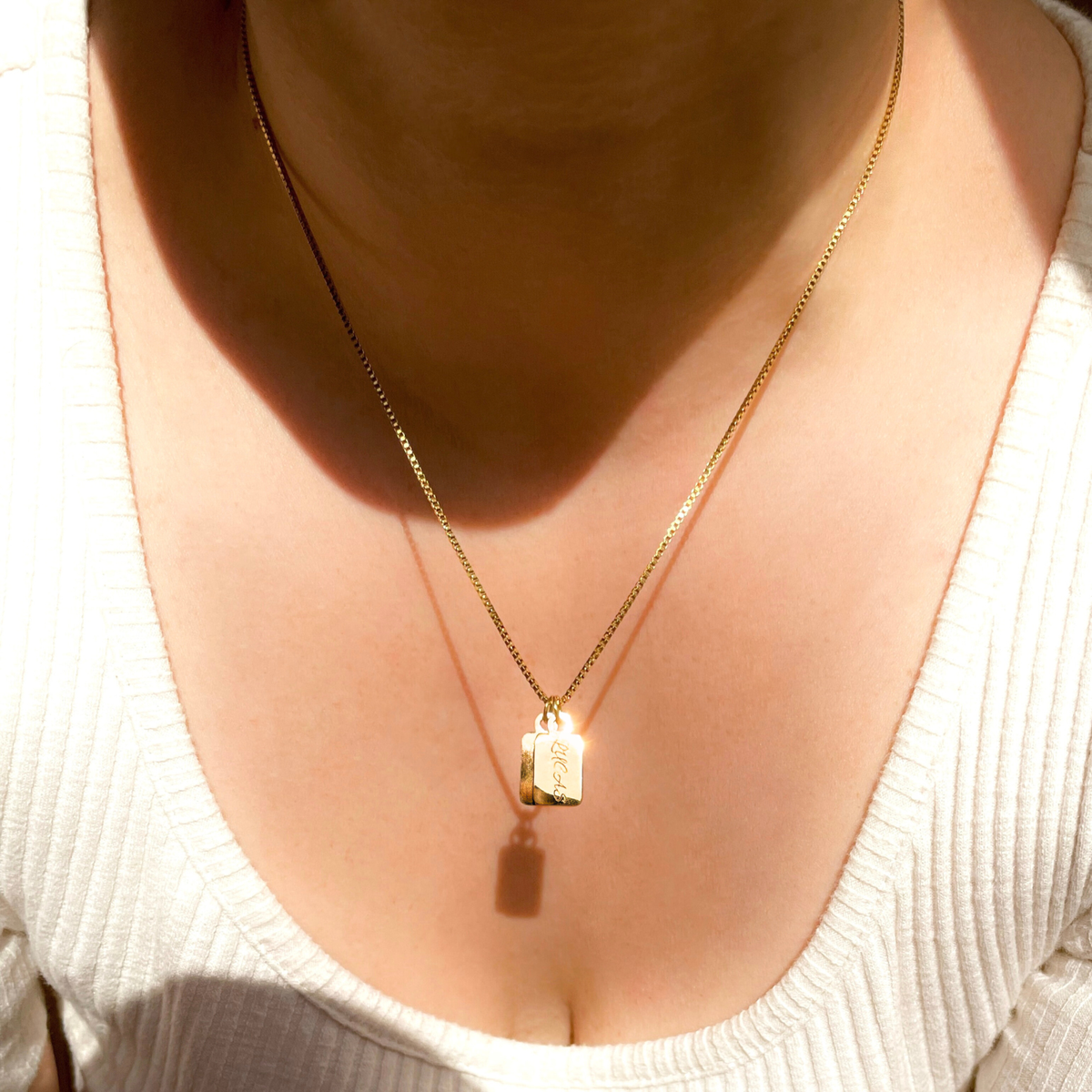 A close-up of a person's neck and chest wearing a delicate gold chain necklace with two small rectangular pendants engraved with names, styled with a white ribbed knit top.