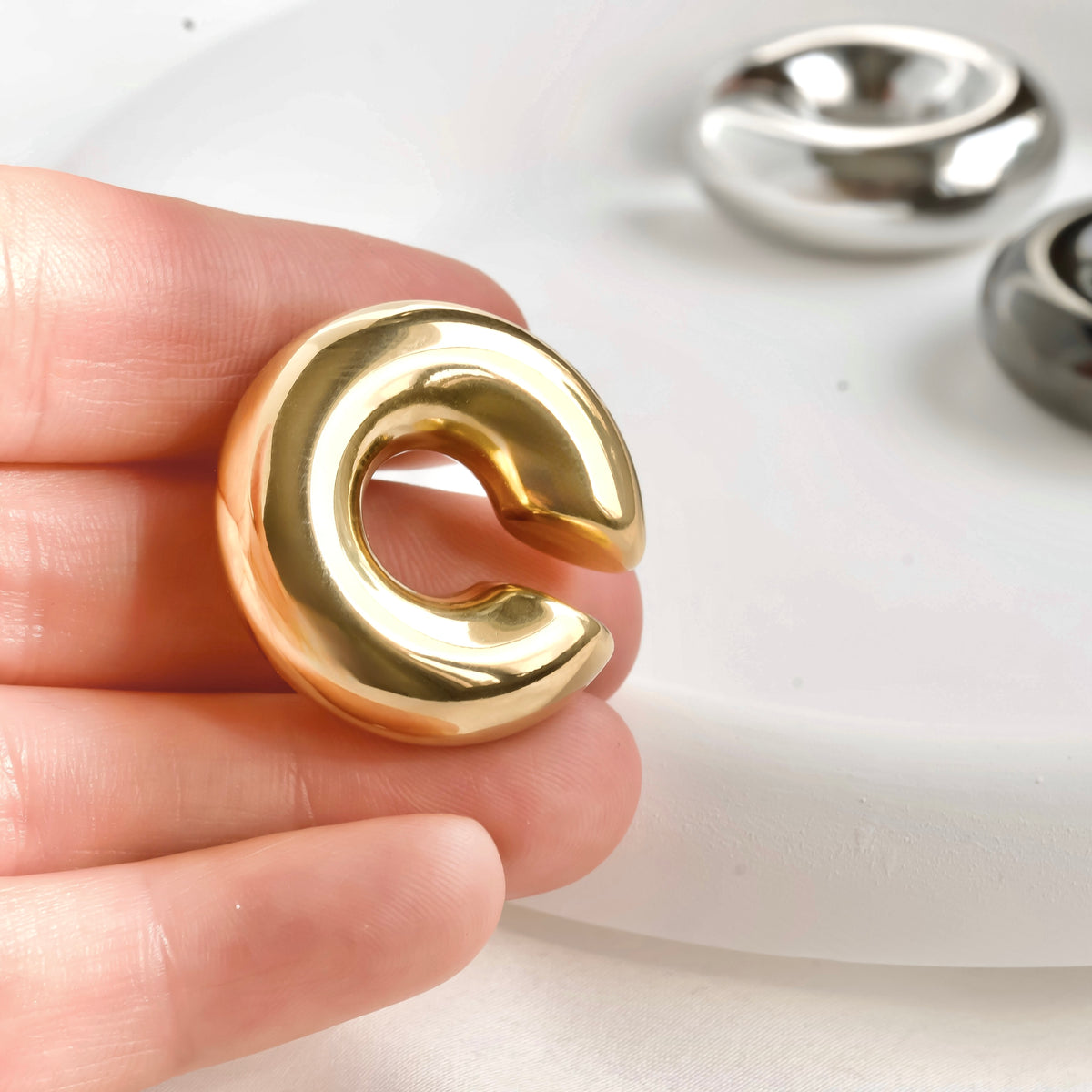 Gold dome ear cuff held between fingers with silver earrings in the background.