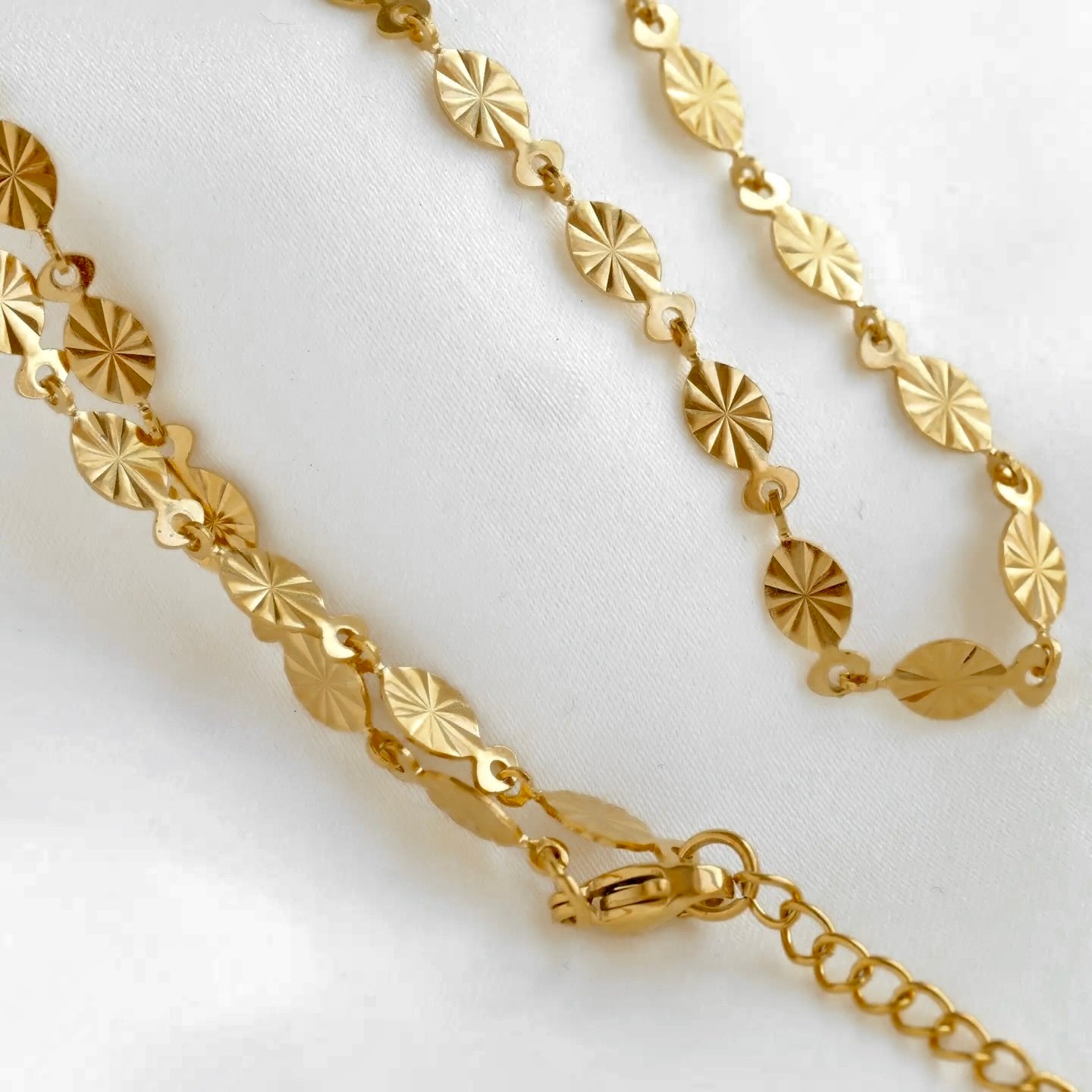 A gold chain necklace with repeating sunburst disc links, displayed on a white fabric background.