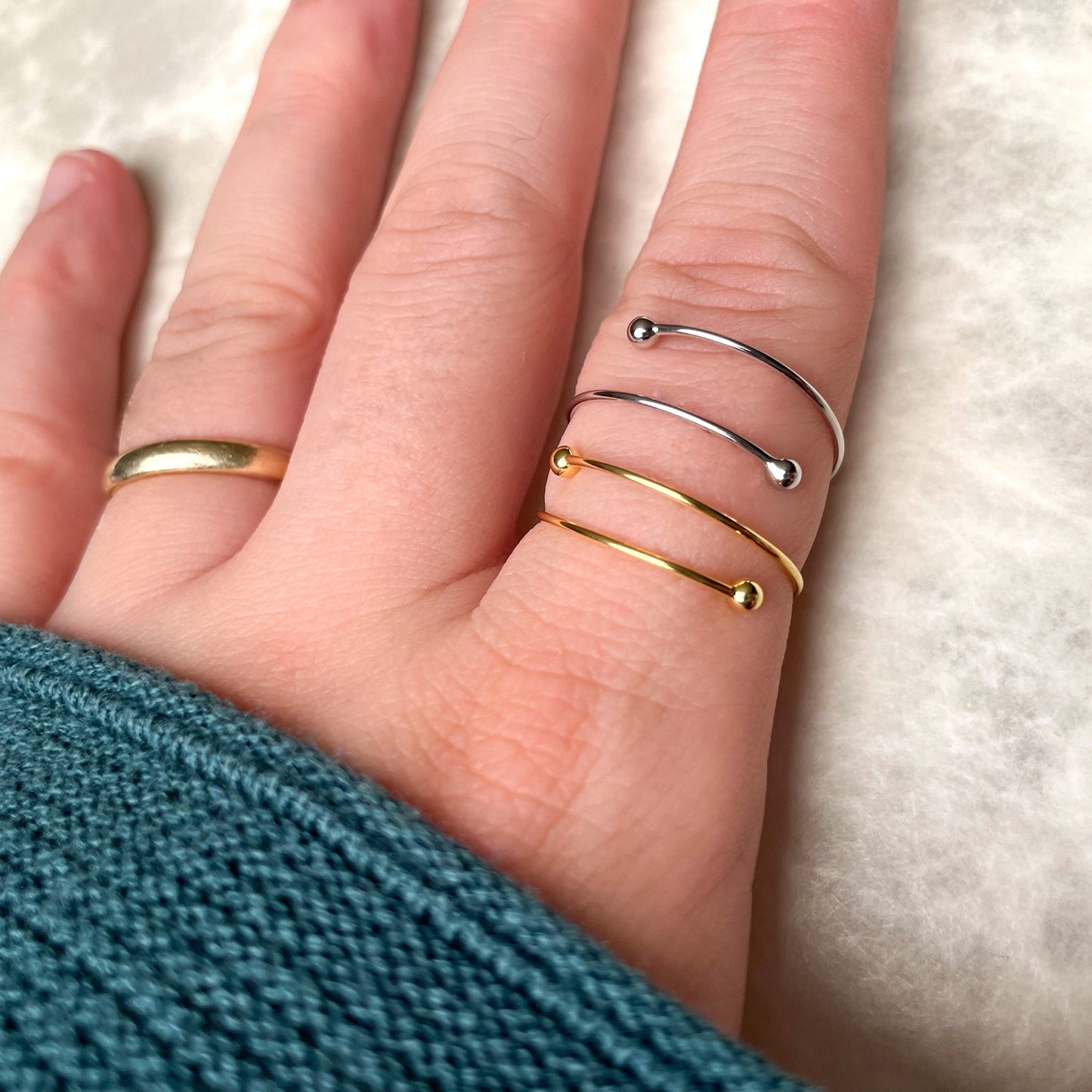 Hand wearing two thin wrap rings, one gold and one silver stacked on index finger, on a light background