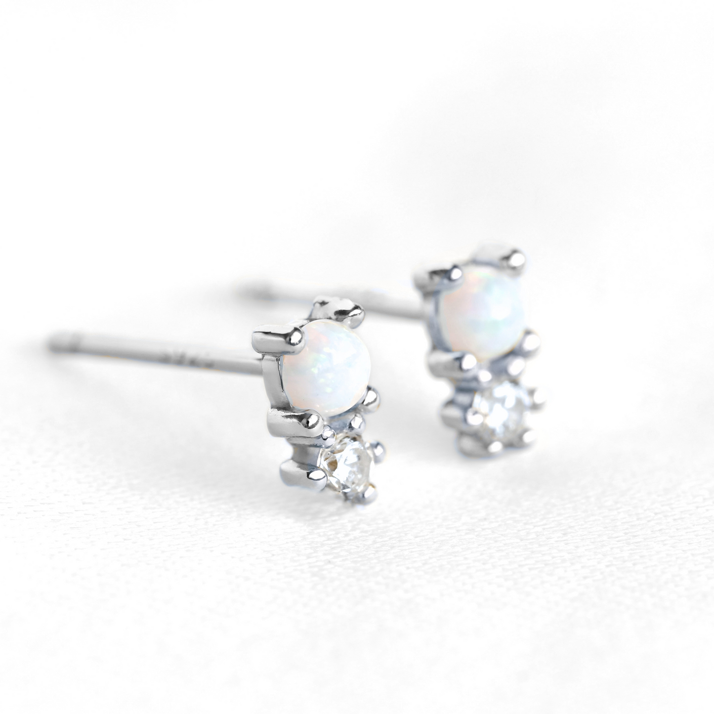 A pair of silver stud earrings featuring a small white opal and a clear cubic zirconia stone, shown on a clean white background.