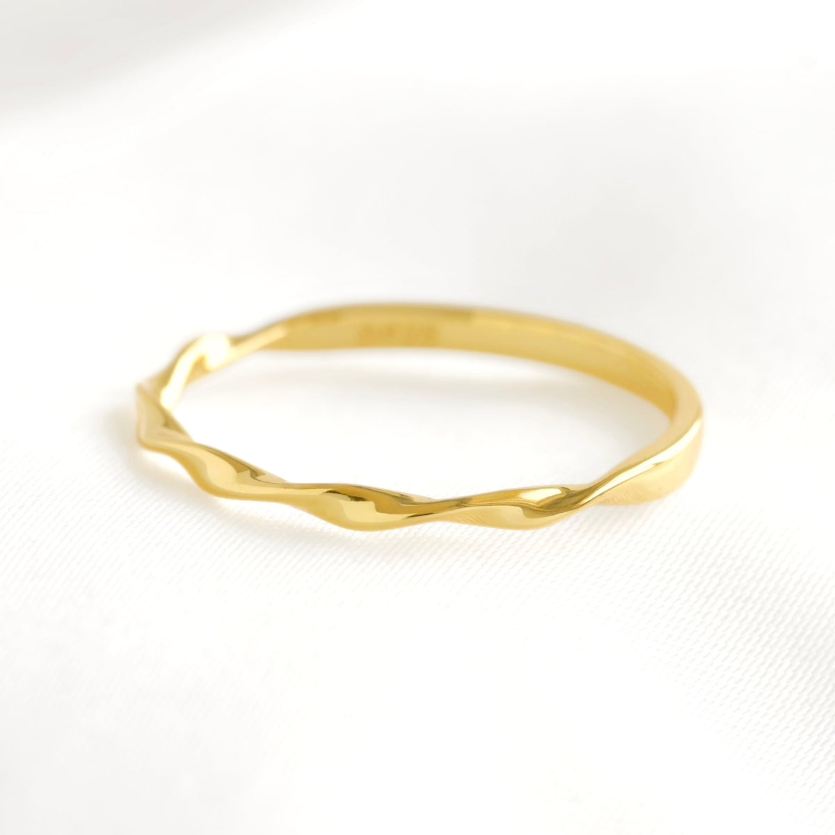 A thin gold ring with a wavy, organic twisted design, photographed on a clean white textured fabric background.