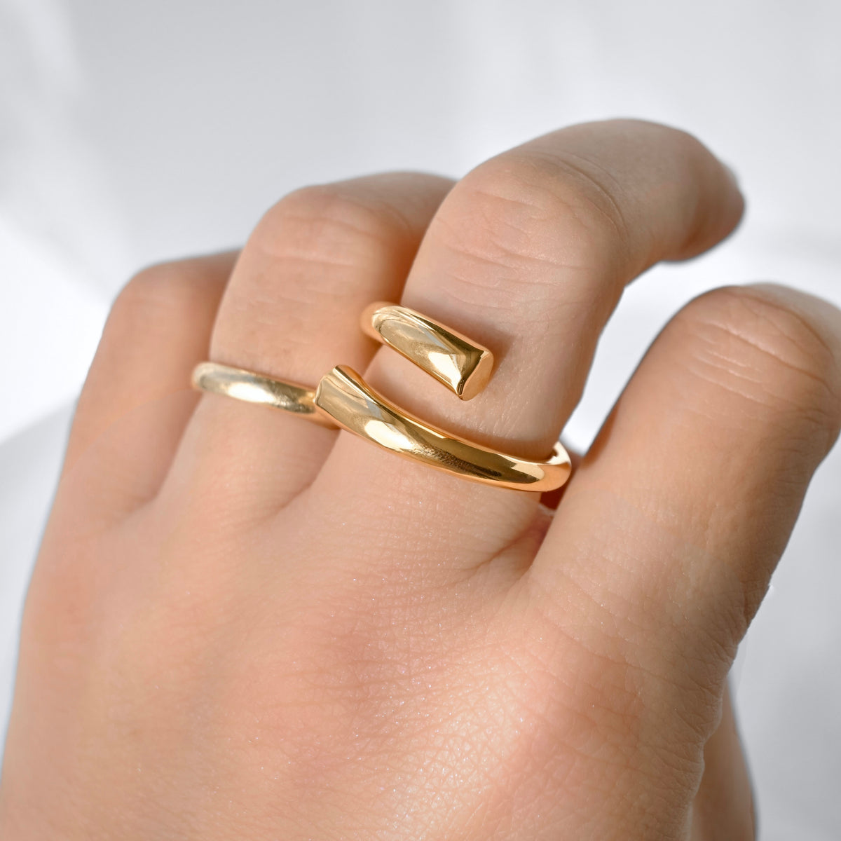 A close-up of a hand wearing a polished gold open wrap ring, showing the smooth metallic band in use on the fingers against a soft white background.