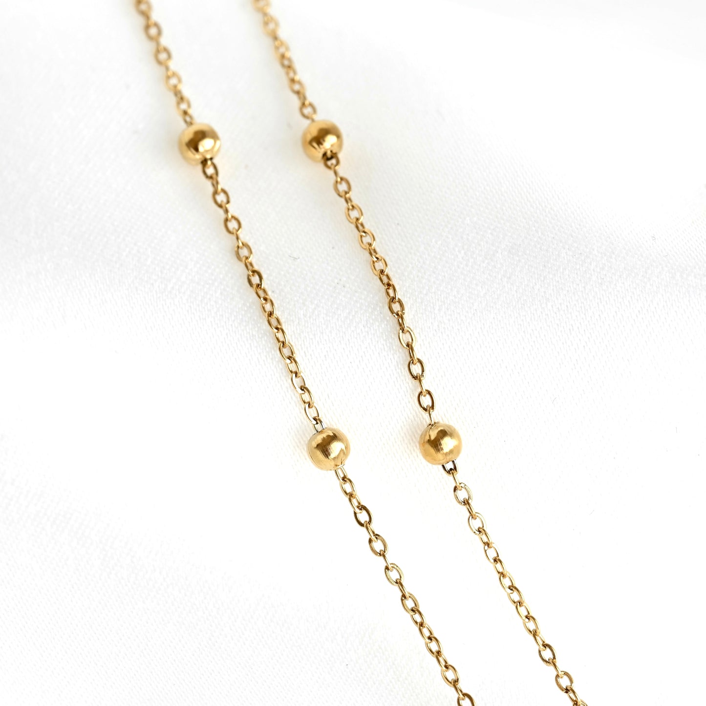A delicate gold chain necklace featuring four evenly spaced, polished round gold beads, displayed on a soft white fabric background.
