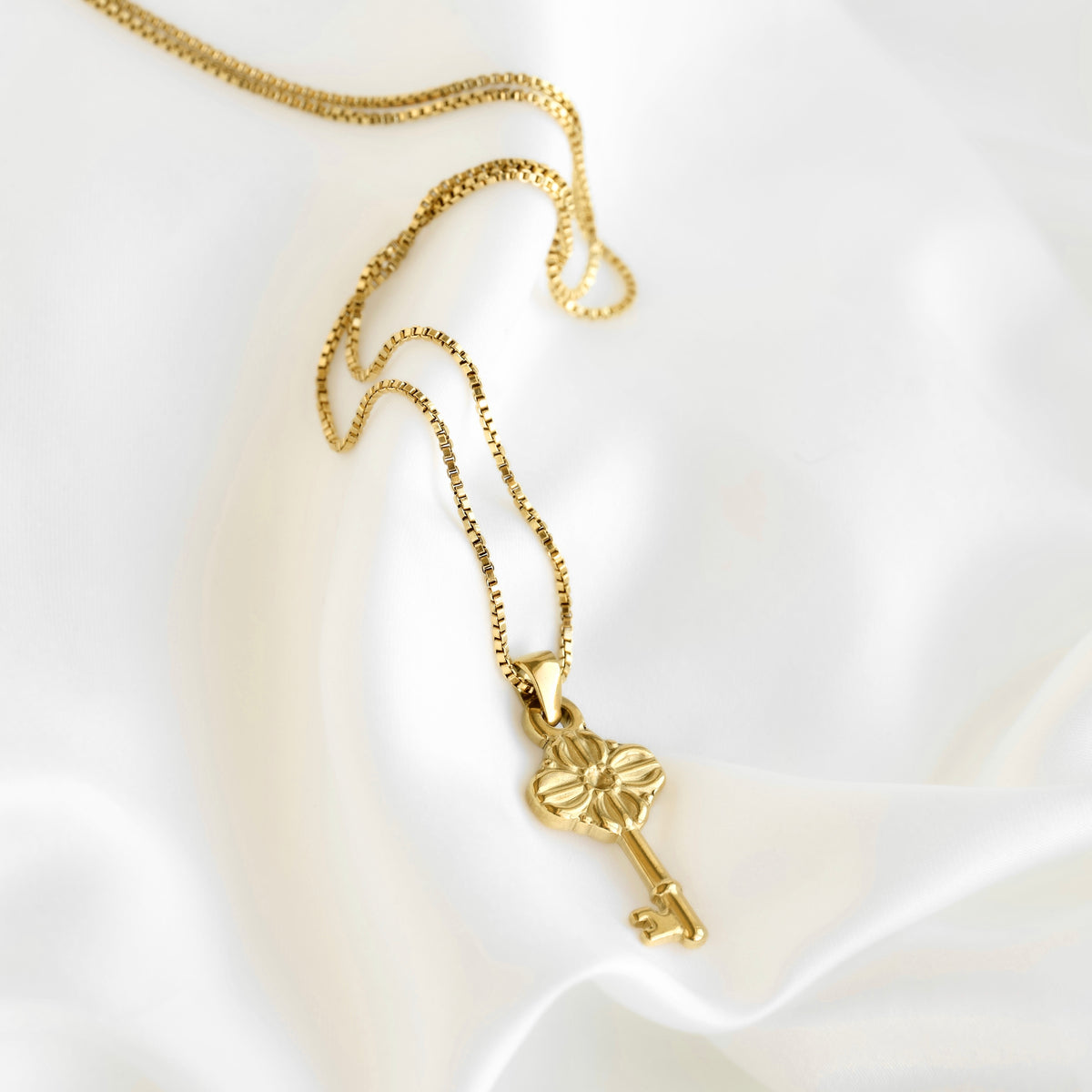 A gold key-shaped pendant necklace with a flower-like cutout detail, resting on draped white satin fabric.