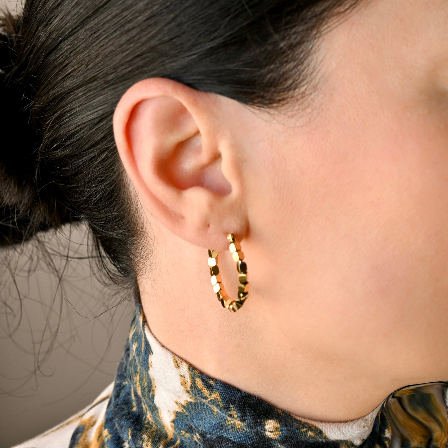 A close-up of a person's ear wearing a textured gold hoop earring, with dark hair pulled back and a patterned blue and gold top visible.