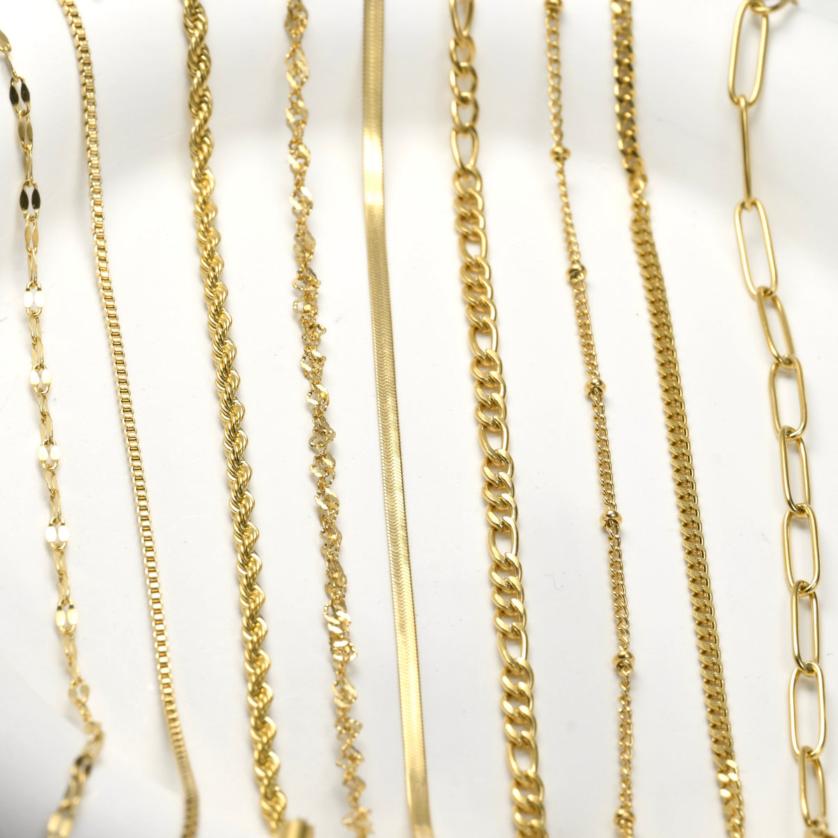 Collection of various dainty gold chain bracelets on a white background. The designs are as follows from left to right: dainty sequin, box, rope, singapore, 2mm herringbone, figaro, satellite, curb and thin paperclip