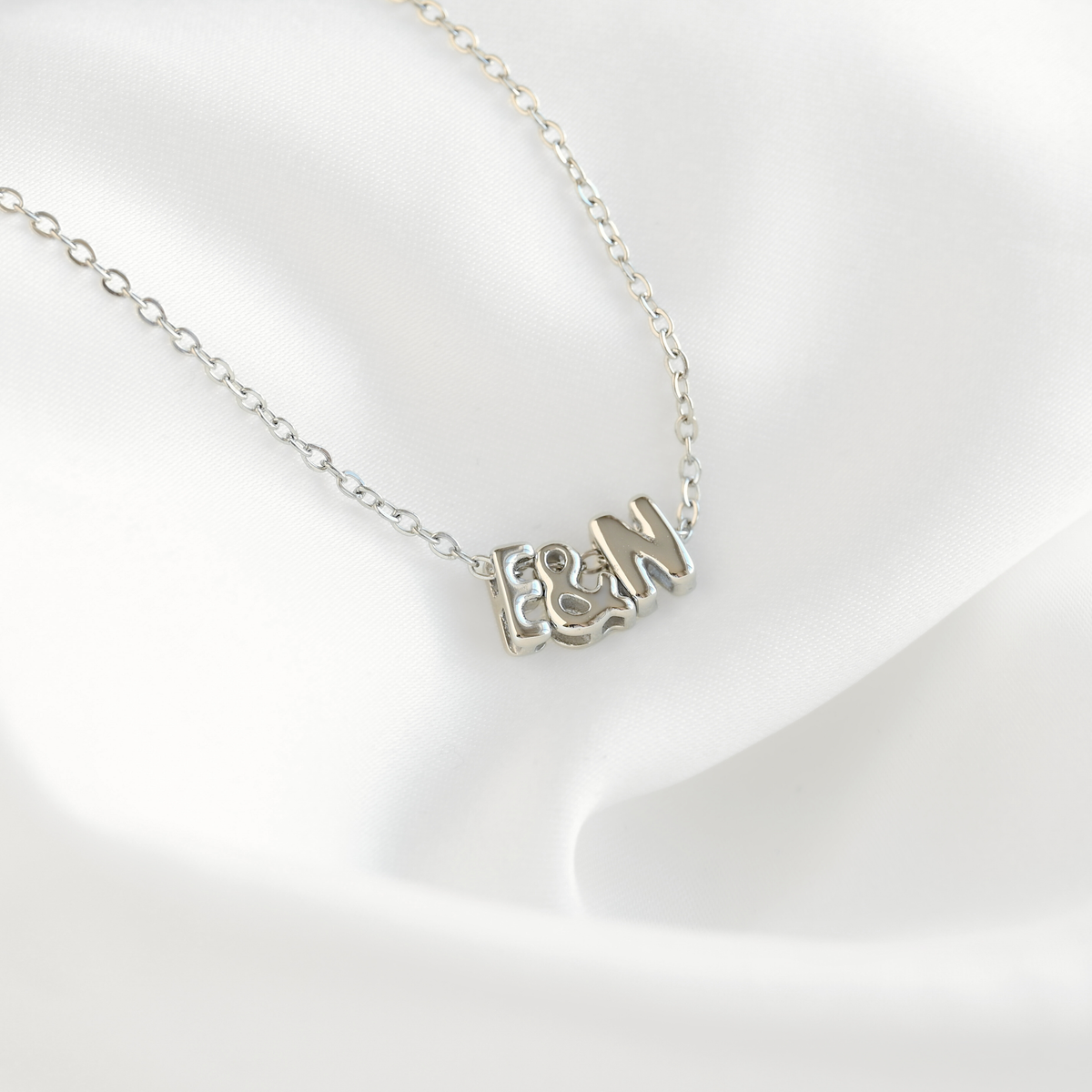 A silver initial necklace featuring the letters 'F', '&', and 'M' in a textured, openwork design, displayed on a soft white satin fabric background.