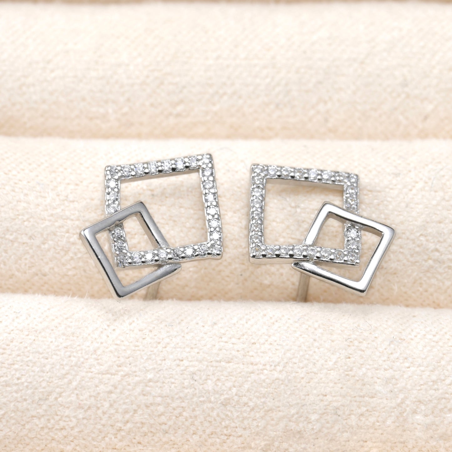 Pair of silver square-shaped earrings with clear stones on a beige fabric background