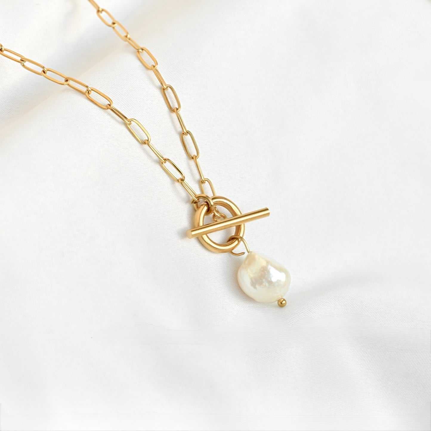 A gold chain necklace featuring a toggle clasp and a dangling baroque pearl pendant, displayed on a soft white fabric background.