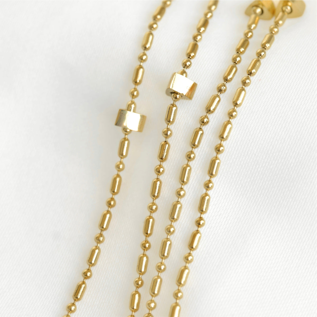 A close-up of a gold-toned beaded necklace laid on a white fabric background, showing elongated oval beads, small round beads, and two small rectangular spacer details.