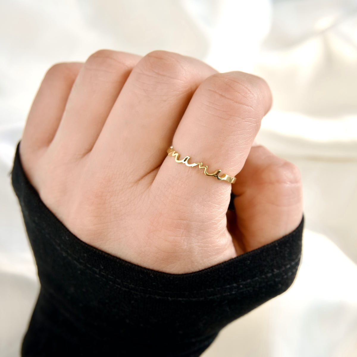 A close-up of a hand wearing a delicate gold ring with a wavy script 'mama' design, set against a soft white background.
