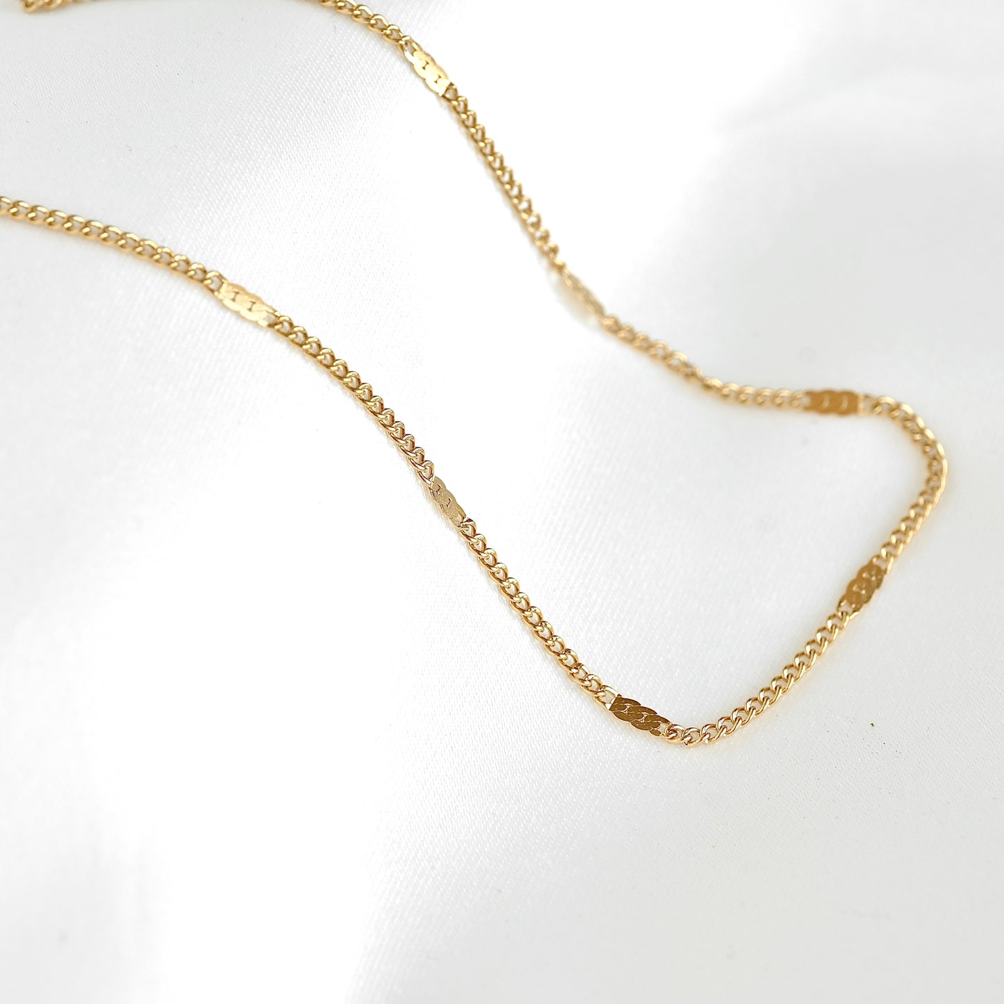 A delicate gold chain necklace with small rectangular links, laid out on a white fabric background.