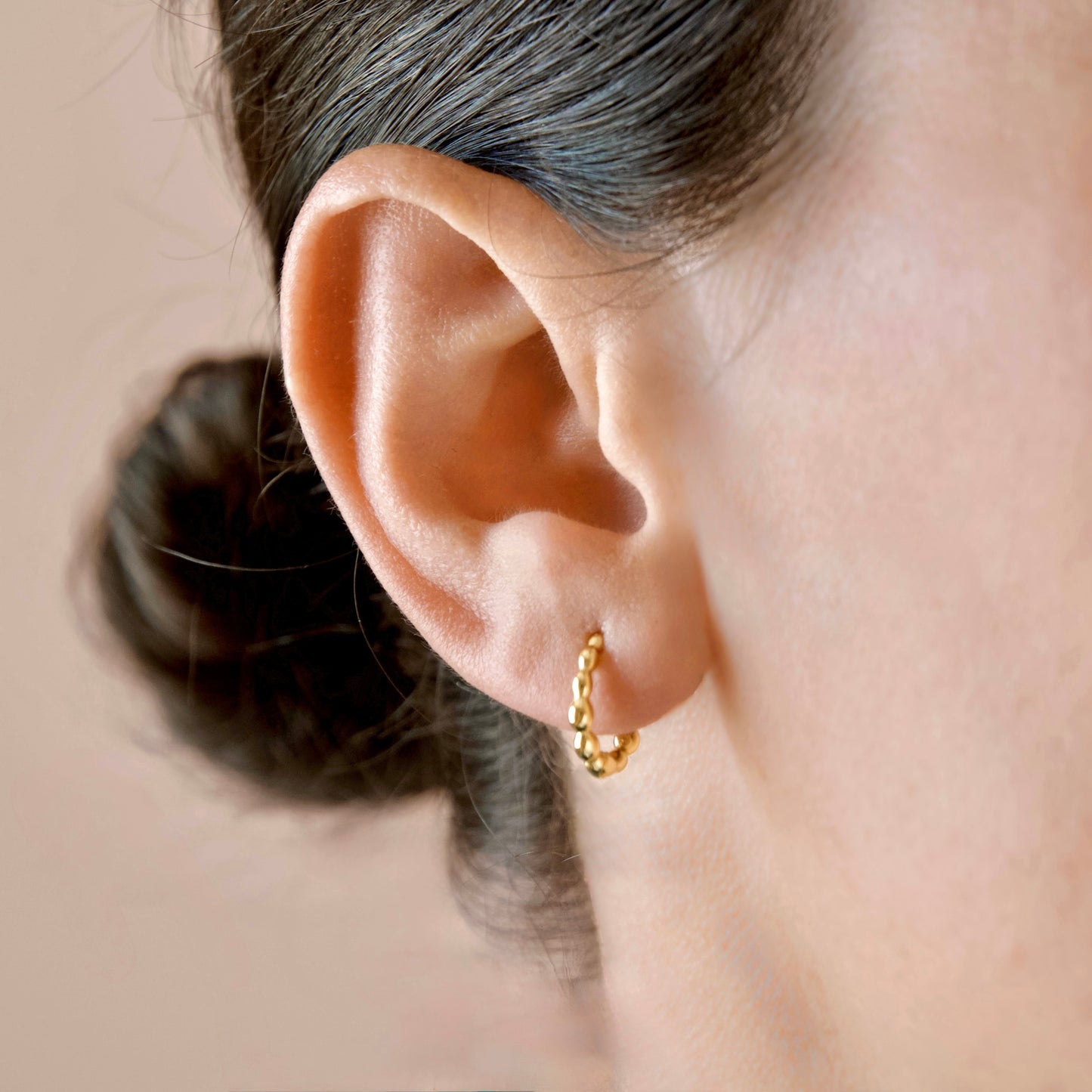 A close-up of a person's ear wearing a small gold huggie hoop earring against a soft beige background.