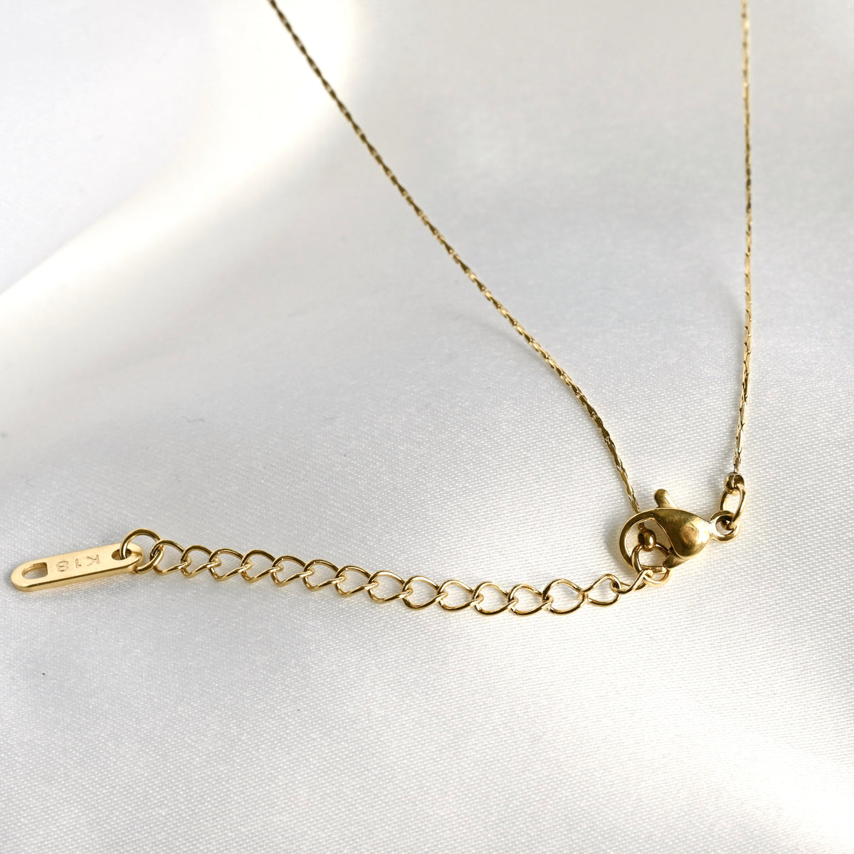 Close up view of the gold solitaire necklace's lobster claw clasp, extender and rectangular 'K18' plating hallmark tag on a white background