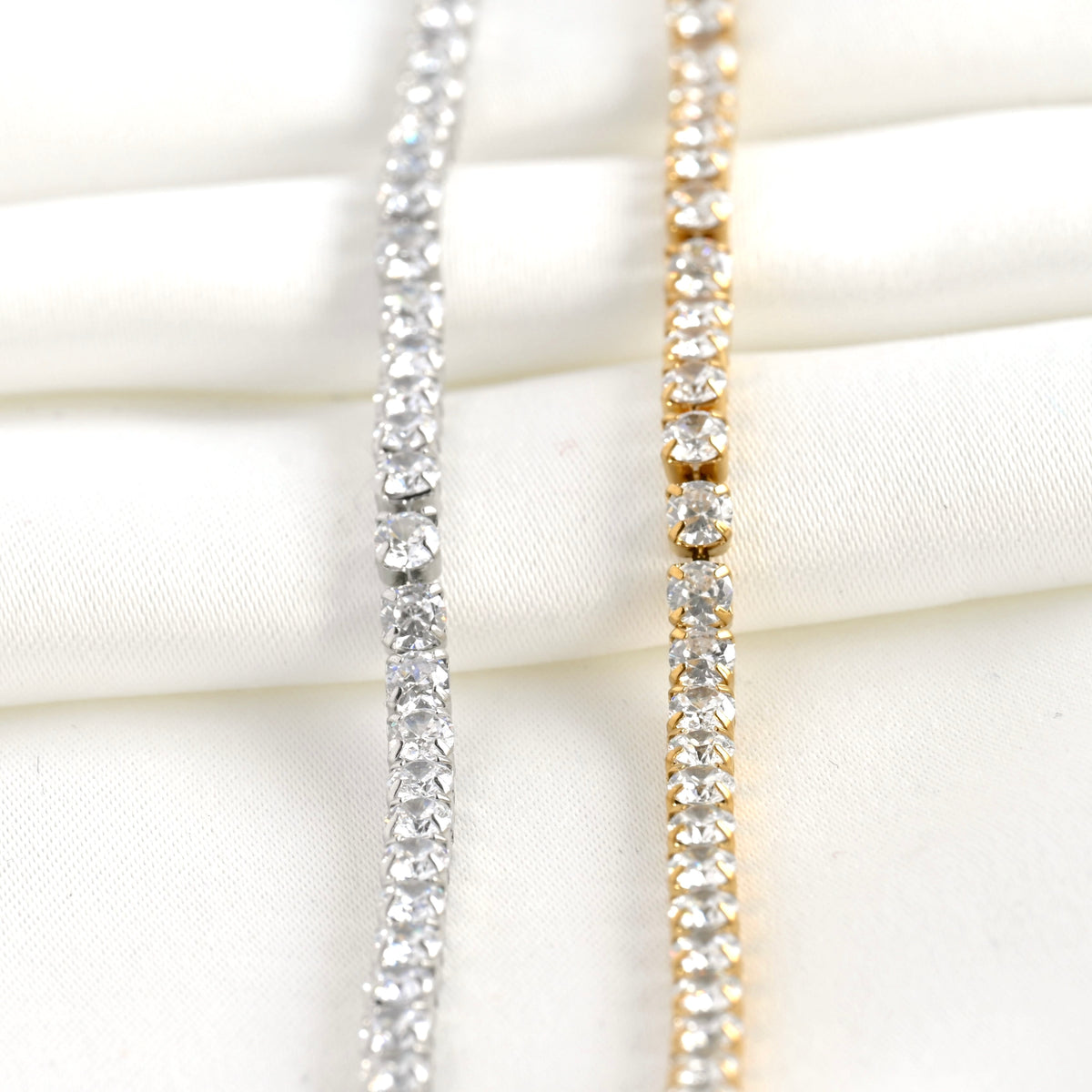 Front vertical view of two diamond tennis chains, one silver and one gold, on a white fabric background.