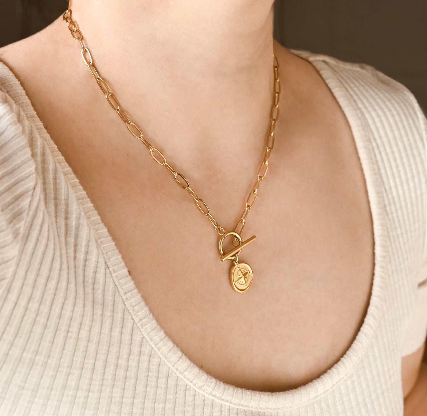 A close-up of a person wearing a gold chain necklace with a toggle clasp and a small oval pendant featuring a star design, styled with a cream ribbed top.