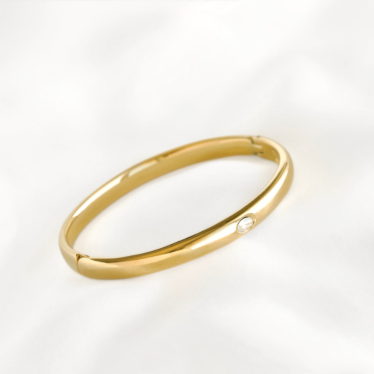A polished gold bangle bracelet with a small round clear gemstone accent, displayed on a soft white fabric background.