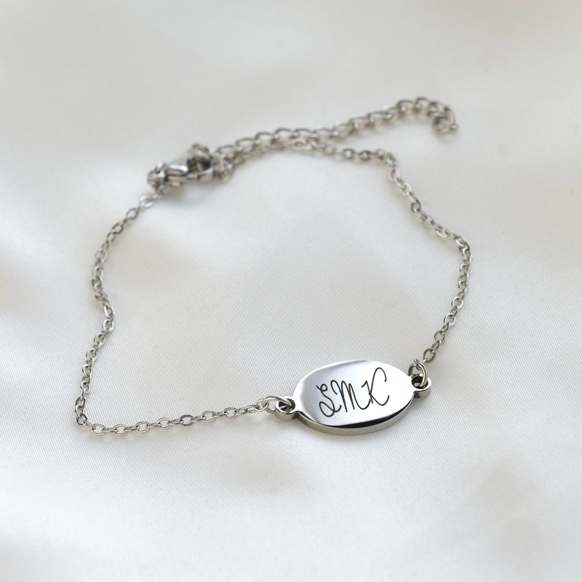 A delicate silver chain bracelet with an oval engraved plate featuring the initials 'SMX', displayed on a soft white fabric background.