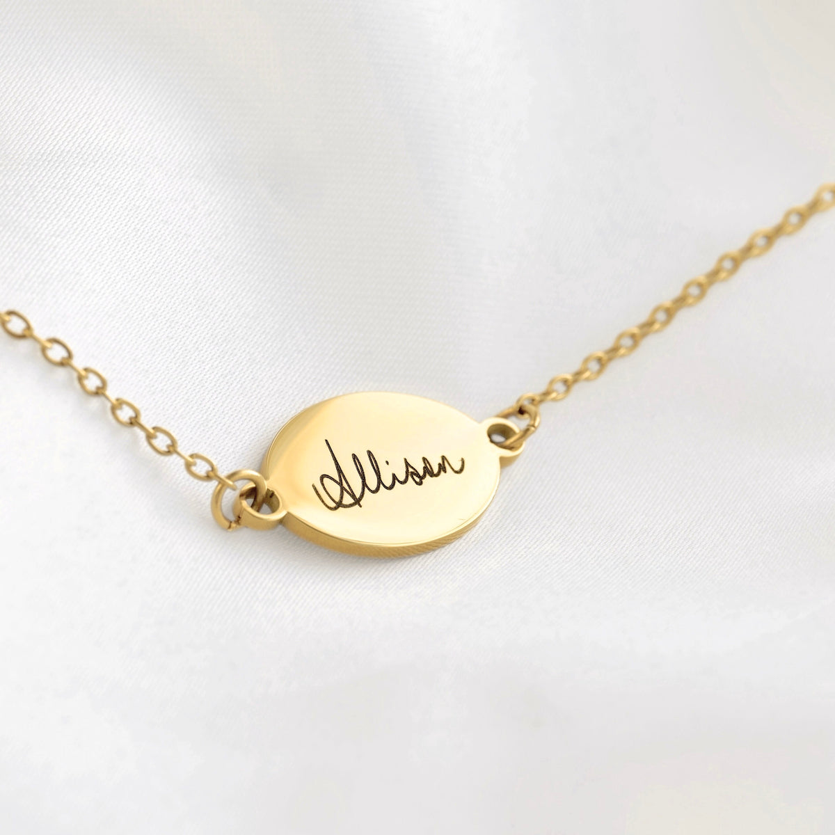 A gold oval nameplate bracelet engraved with the name 'Allison' in cursive, resting on a white satin fabric background.
