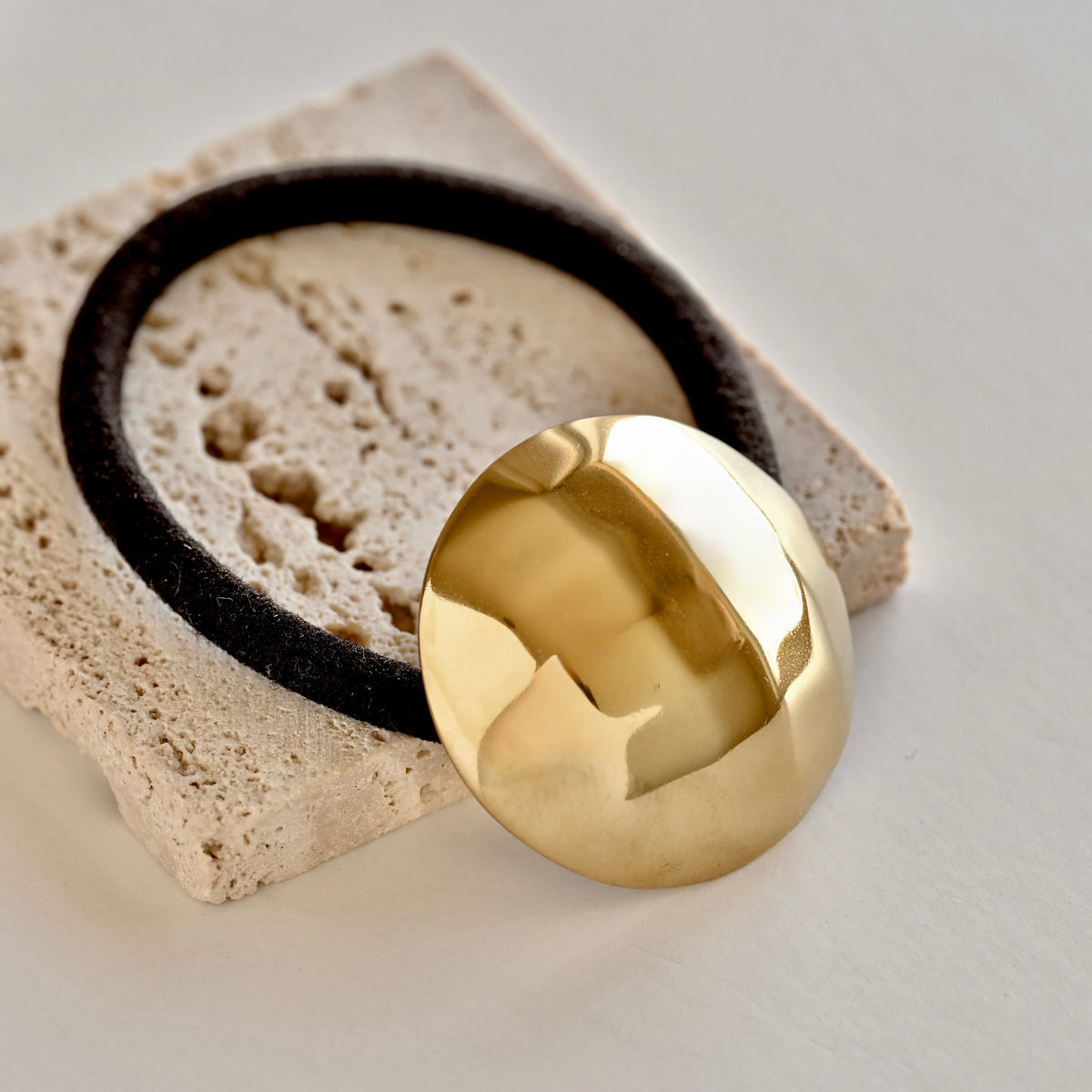A polished, highly reflective gold circular disc or medallion resting on a textured beige stone tile, with a black circular ring partially visible behind it.