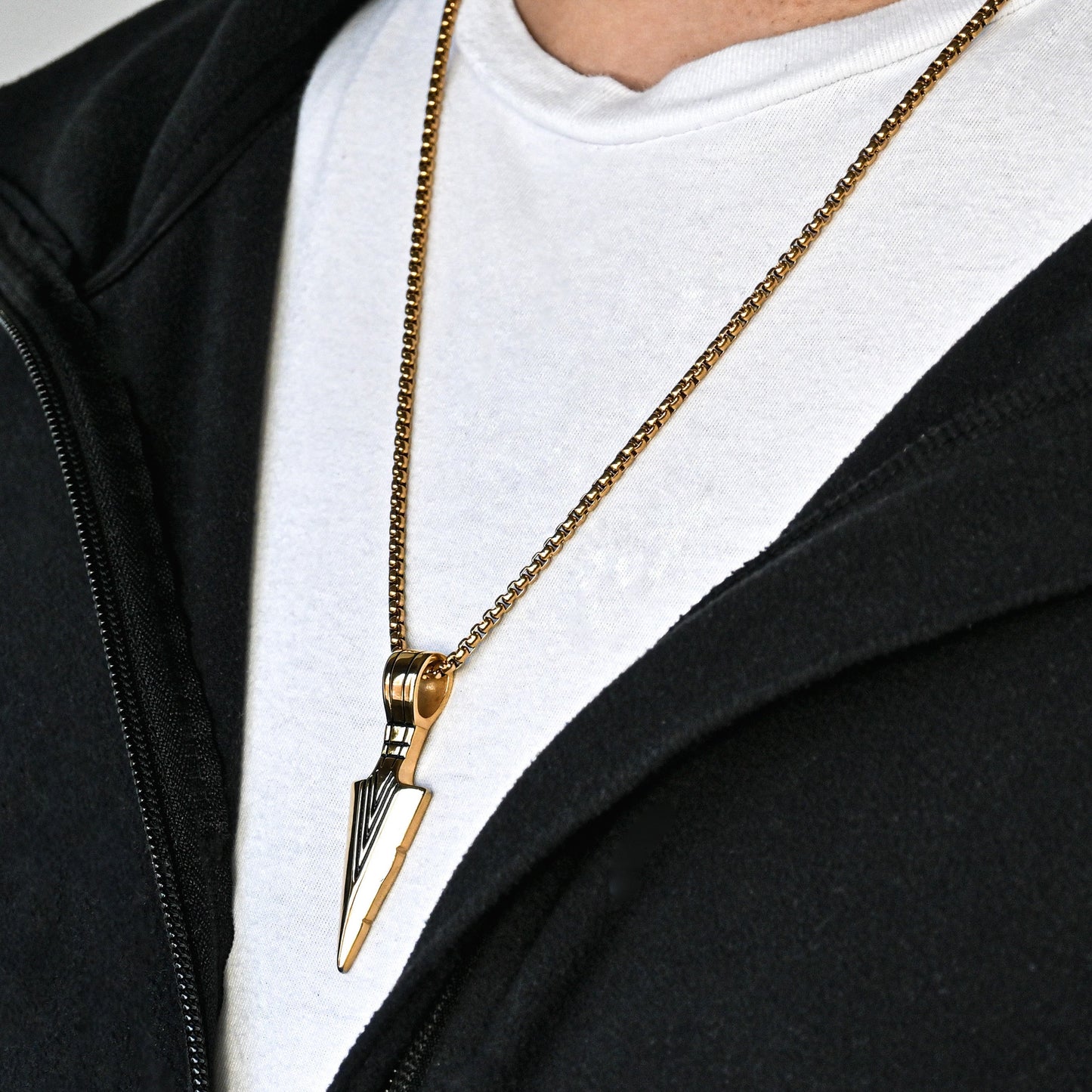 Side view of man wearing a black jacket with a gold necklace featuring an arrowhead pendant on a plain background