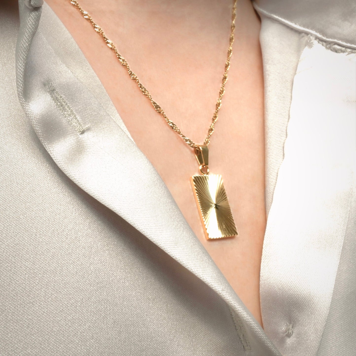 Angled view of radial sunburst necklace with a rectangular pendant on a person wearing off-white satin shirt