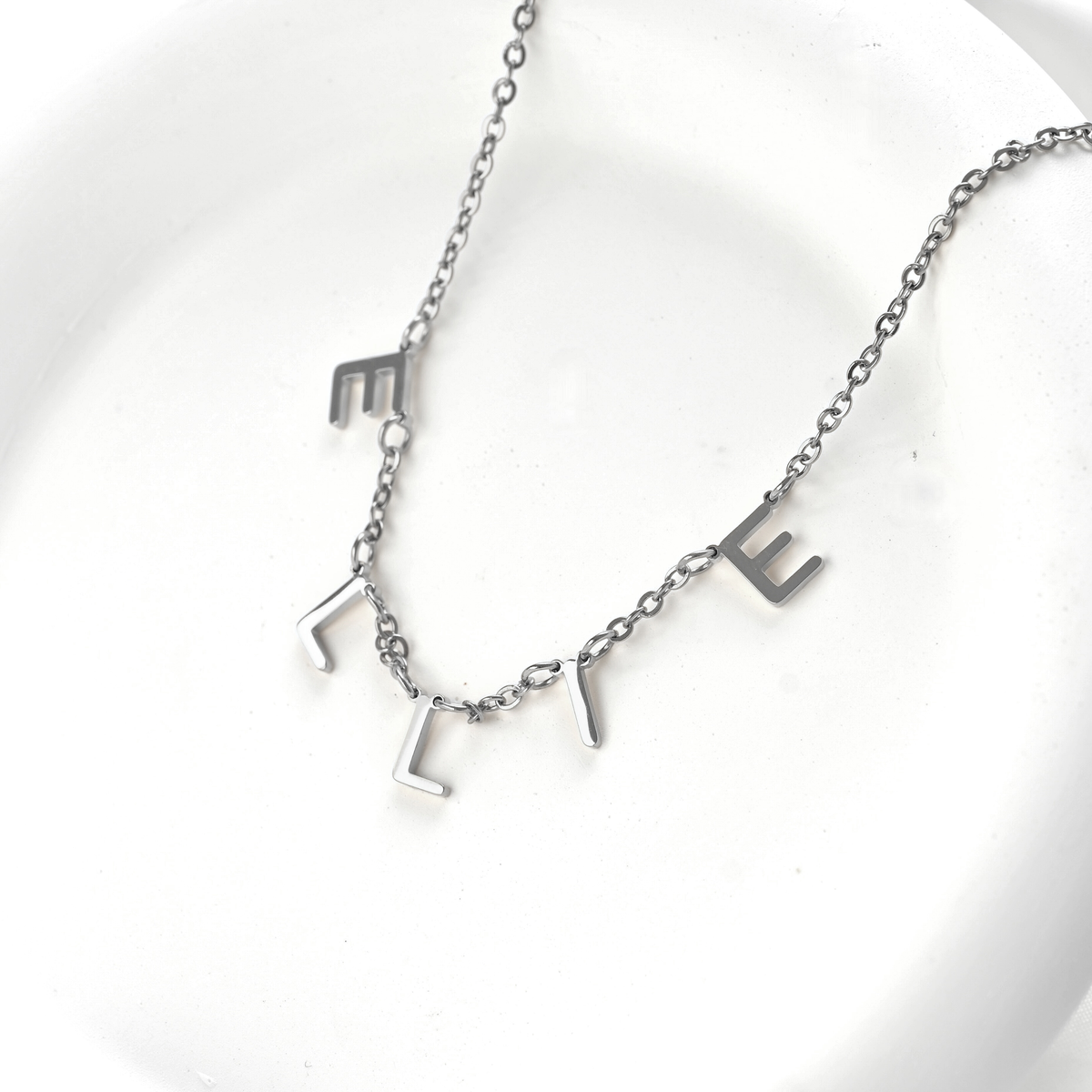 A silver initial necklace spelling 'ELLIE' on a delicate chain, displayed on a white ceramic dish.