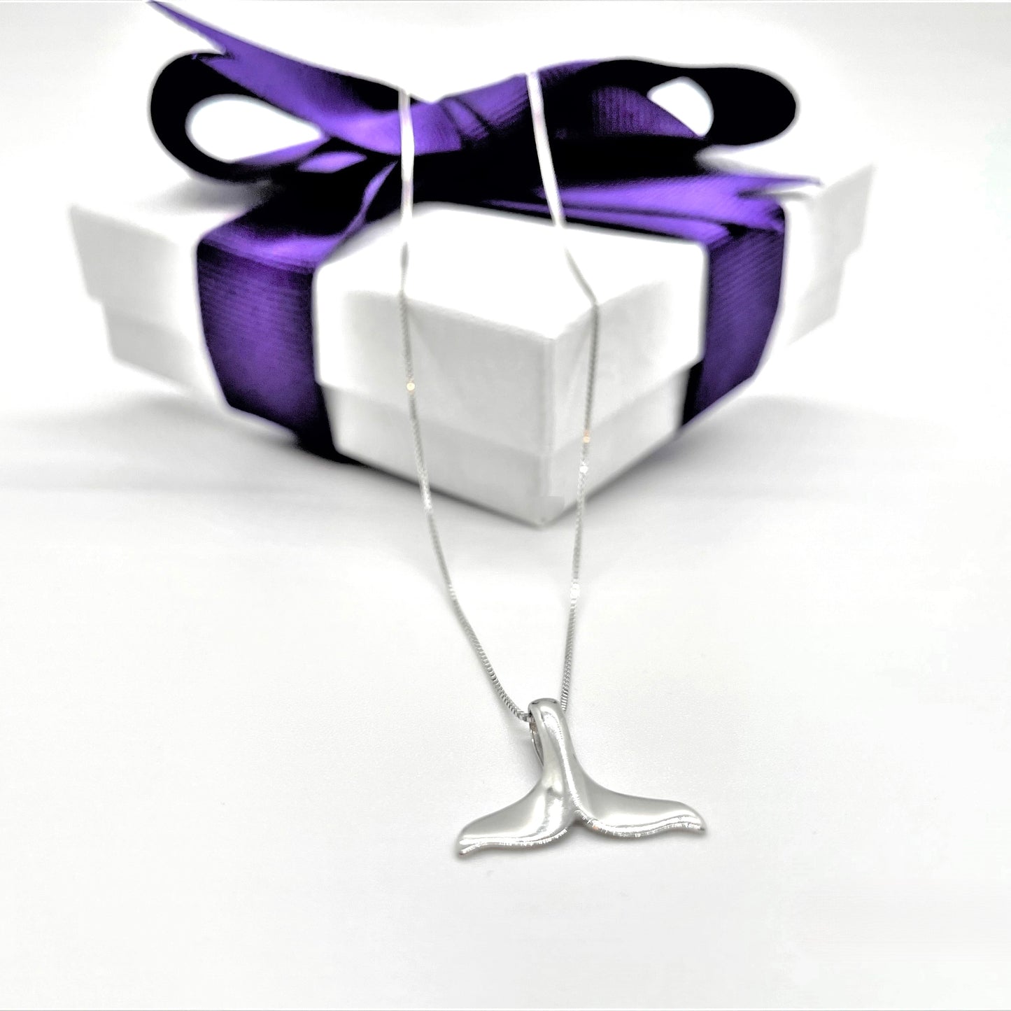 Silver whale tail charm on a white jewelry box with a purple ribbon
