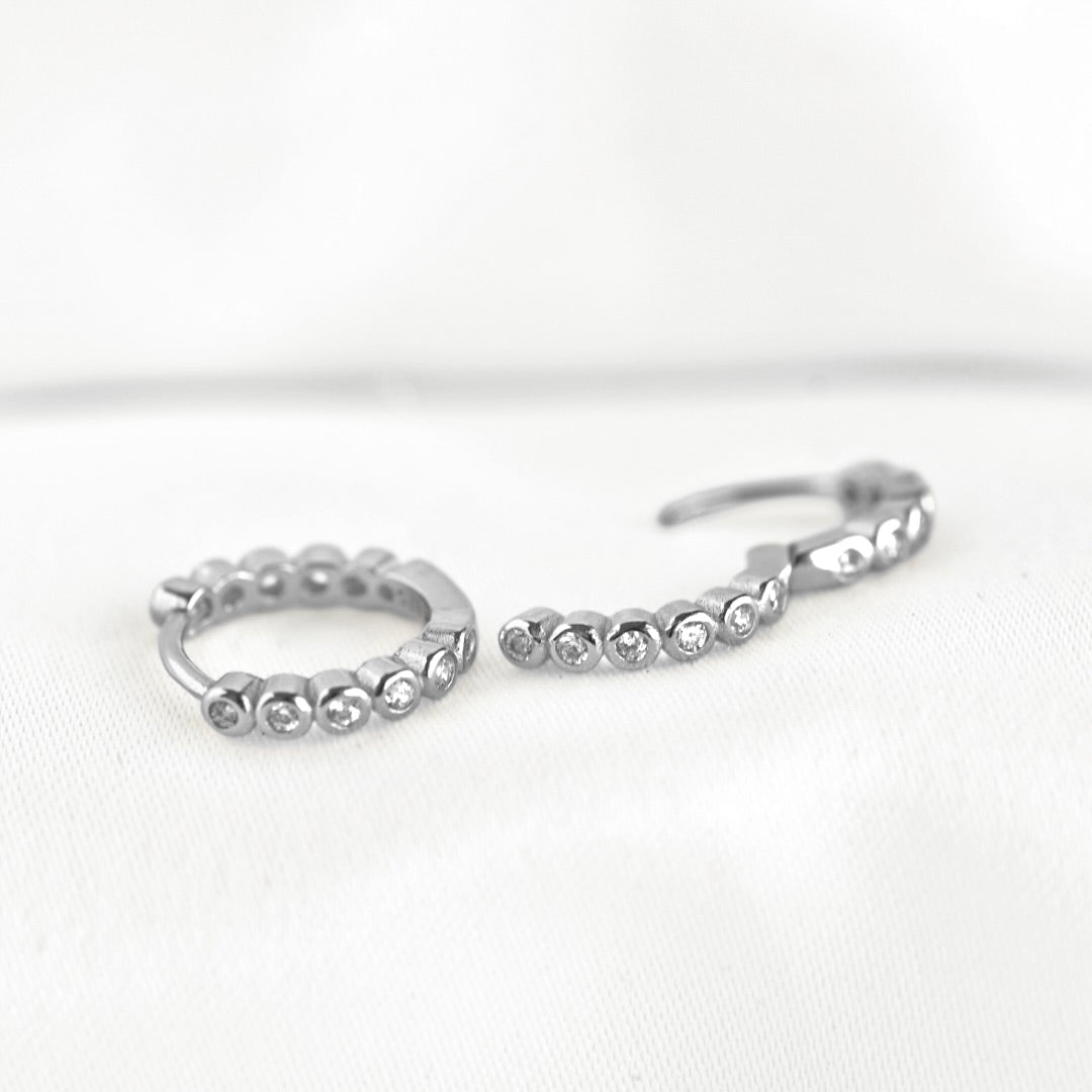 A pair of silver huggie hoop earrings adorned with small clear round stones in a bezel setting, displayed on a soft white fabric background.