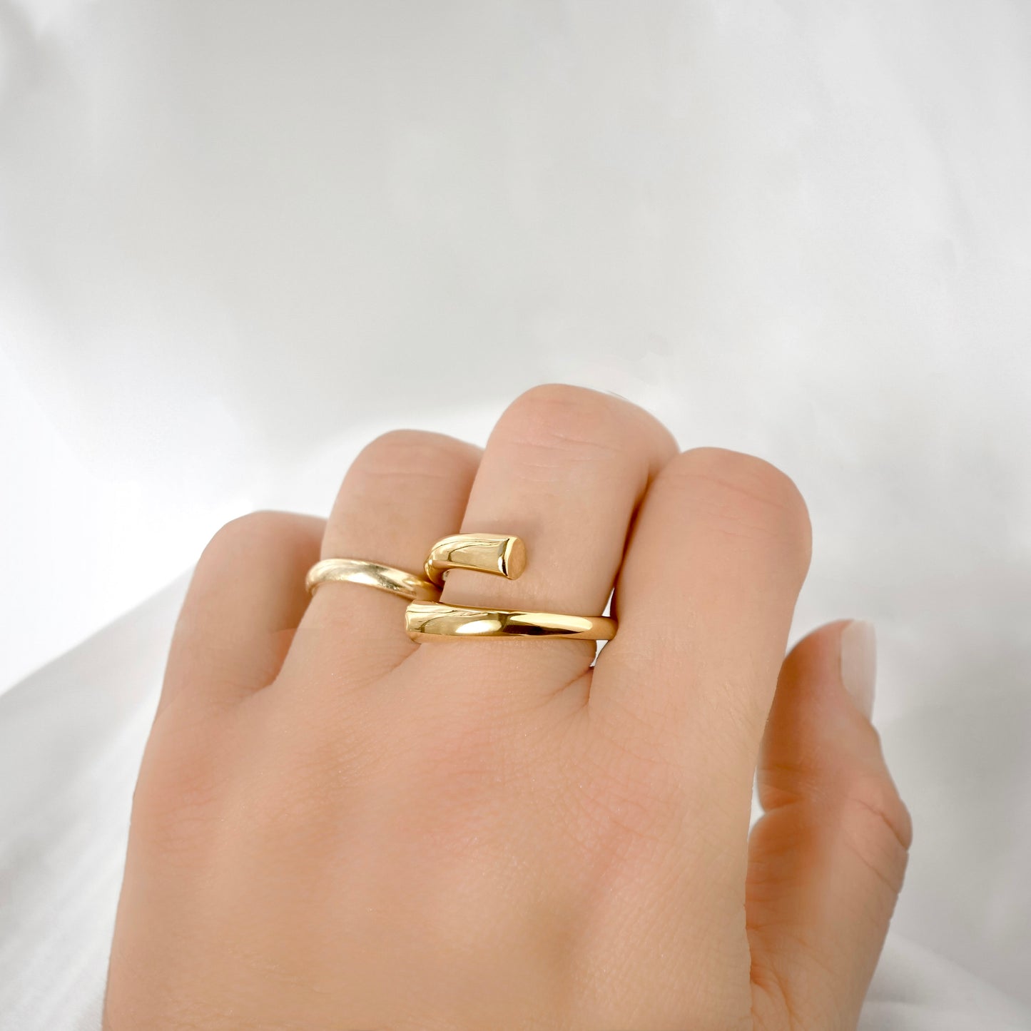 A close-up of a hand wearing two slim gold rings with textured, slightly offset bands against a soft white background.