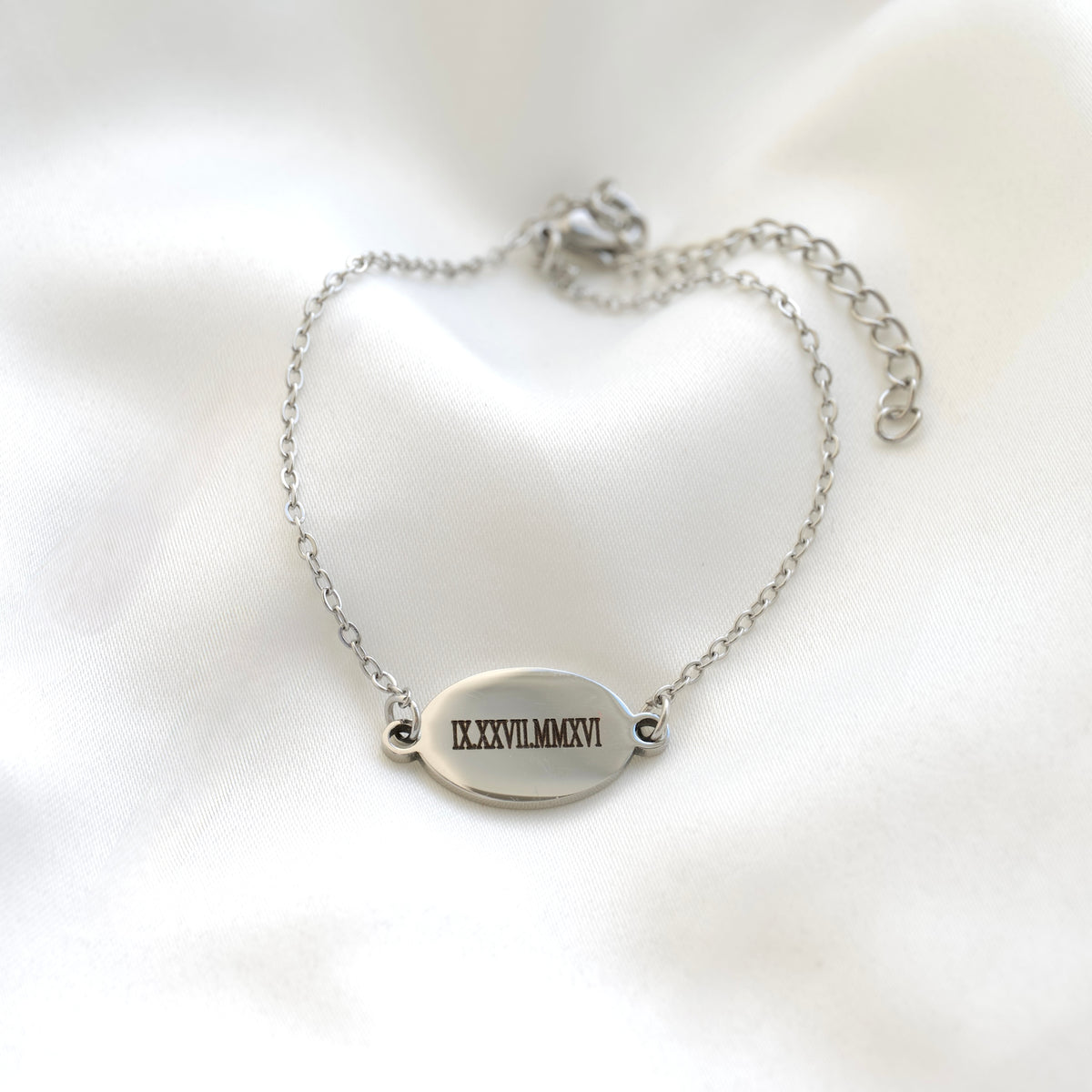 A silver chain bracelet with an oval engraved plate featuring the Roman numerals 'IXXXVIIIMMMXVI', displayed on a soft white satin fabric background.