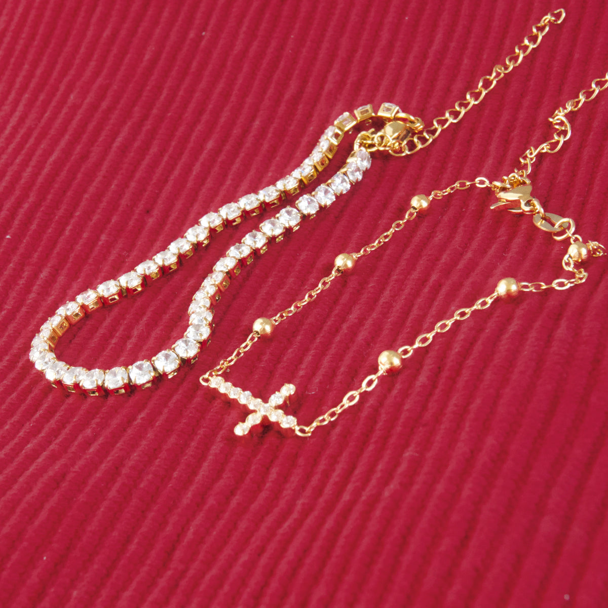 A gold-toned tennis bracelet and matching chain bracelet with a small cross pendant, displayed on a textured red background.