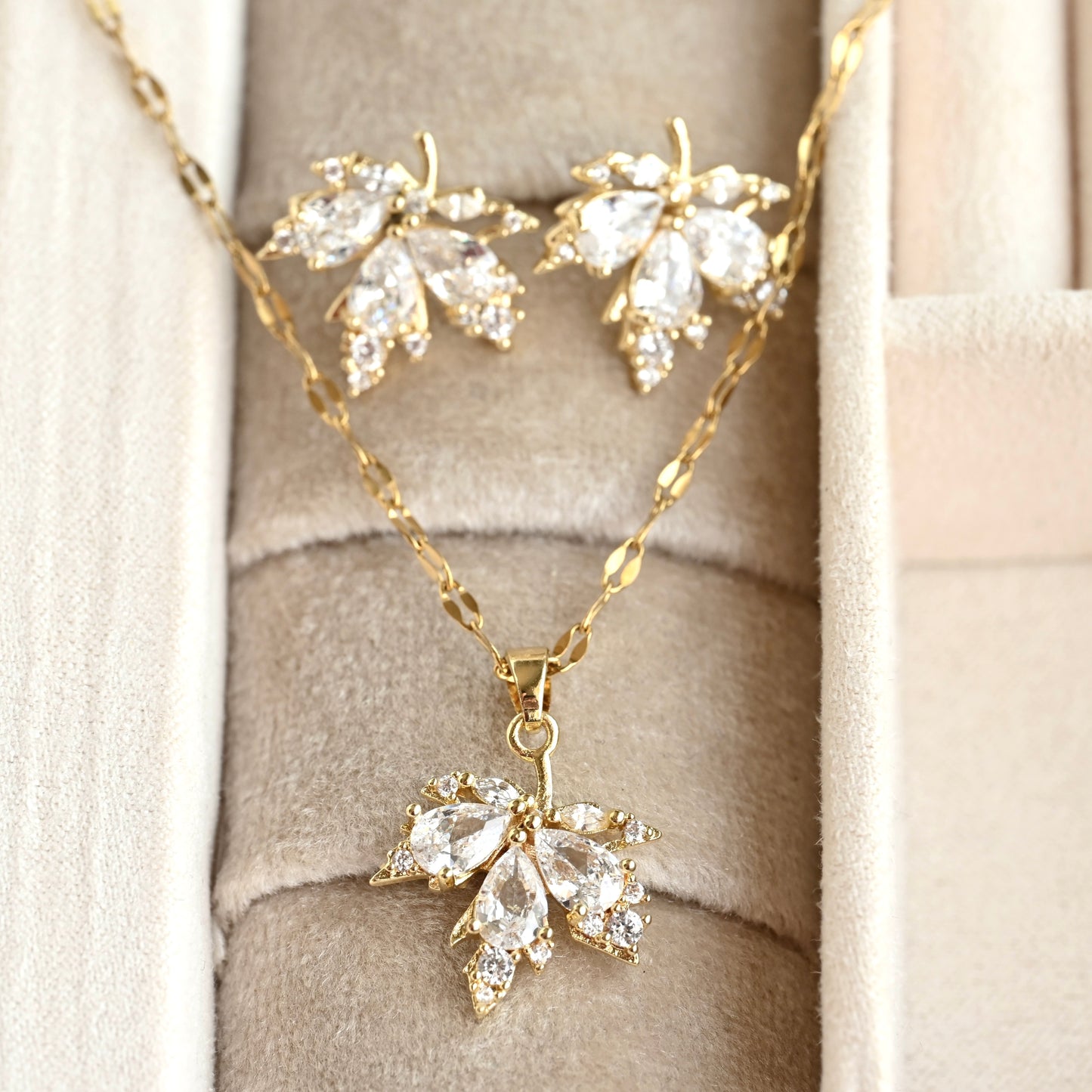 Gold-tone leaf-shaped pendant necklace and matching stud earrings with clear teardrop gemstones, displayed on a beige jewelry box.