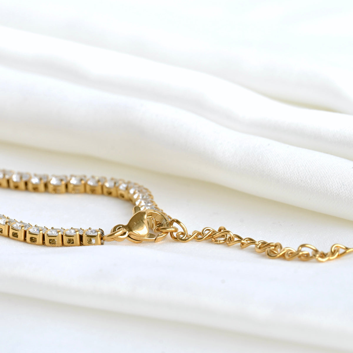 Gold tennis bracelet, close-up view of its lobster claw clasp and extender chain on a white fabric background