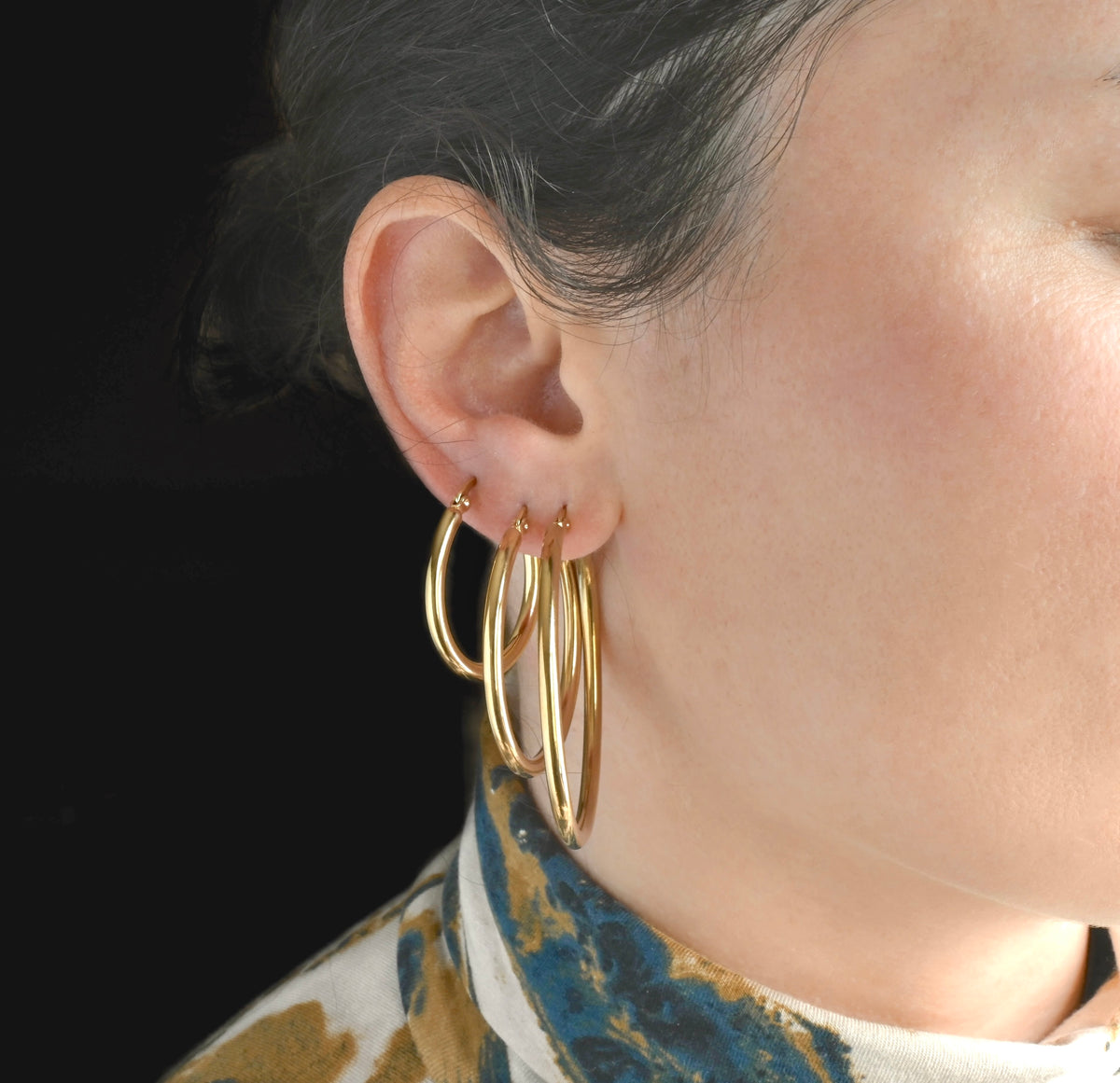 Close-up front view of a person wearing stacked gold tunnel hoop earrings in 30mm, 40mm and 50mm, with a dark background