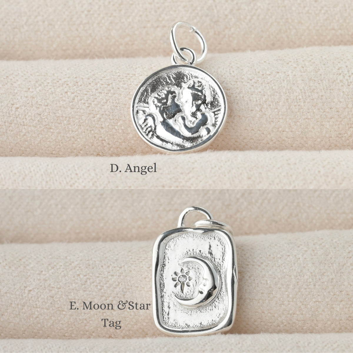 Two silver pendants on a textured beige surface, a coin charm with two embossed angels and an organic tag charm with emmbossed moon and sun/