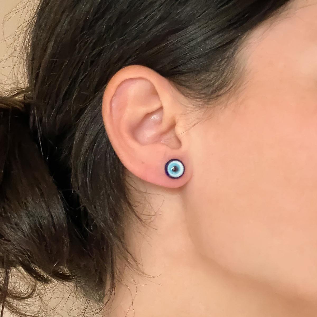 Close-up of an ear with small blue round stud earrings made of glass on a neutral background