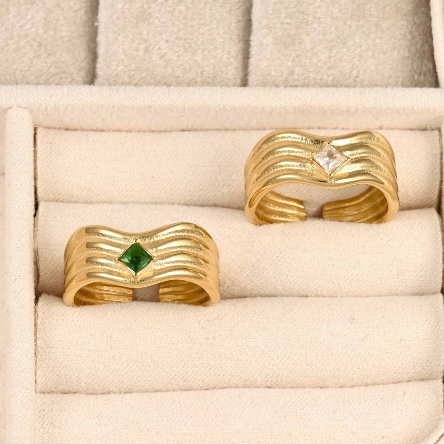 Two gold-colored wavy open-bend rings displayed in a cream-colored velvet jewelry box. One ring features a small green diamond-cut gemstone, and the other features a clear diamond-cut gemstone.