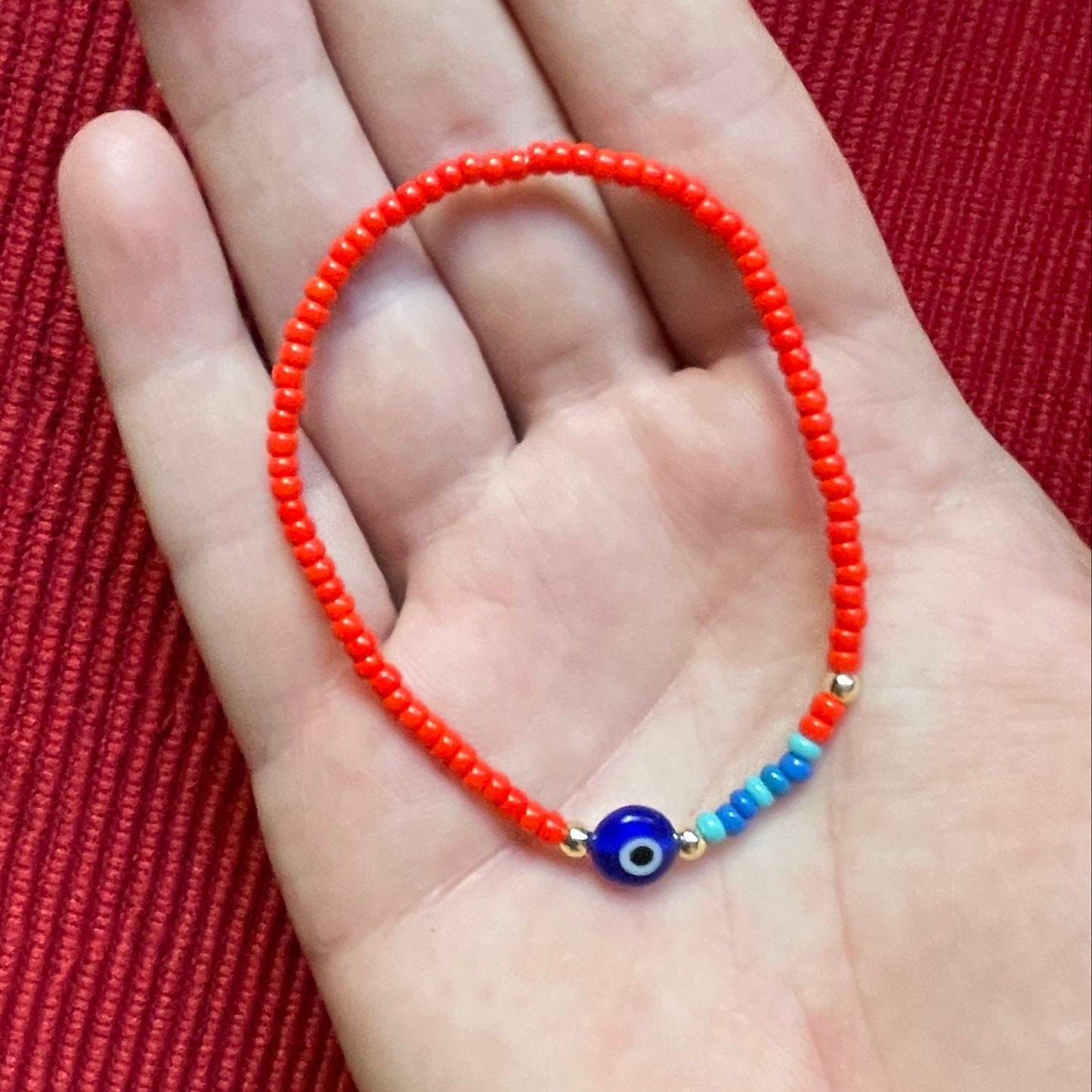 Hand holding a red beaded bracelet with a blue and gold charm on a red textured background